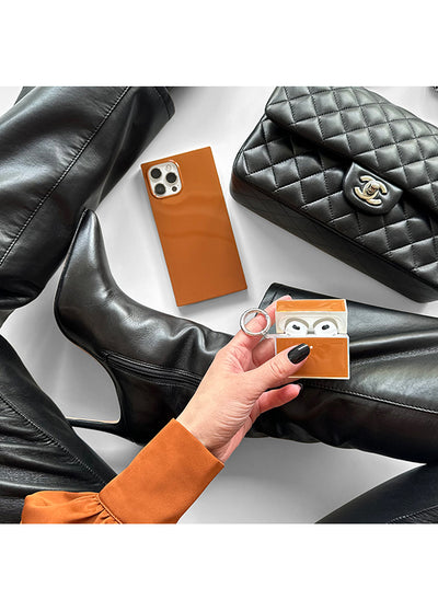 Square caramel brown leather AirPods case held in hand, displayed with black leather accessories on white surface.