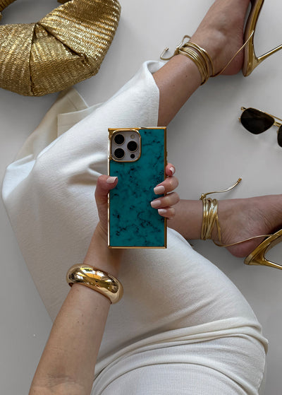 Turquoise iPhone case with gold-tone square frame and matching camera ring, held in hand.