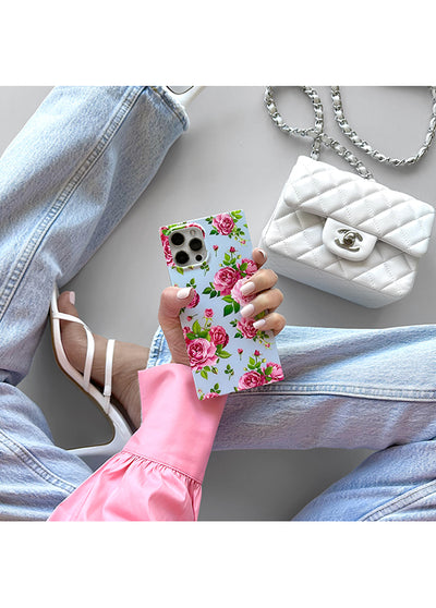 Pink rose floral print iPhone case with square edges held in hand, showing circular camera cutouts on light blue background.