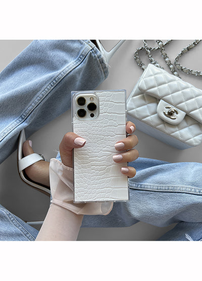 White crocodile-textured phone case with square shape and dual camera cutouts, held in hand next to white quilted bag.