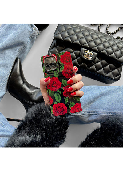 Phone case with red rose print and green leaves on black background, held in hand with red nail polish.