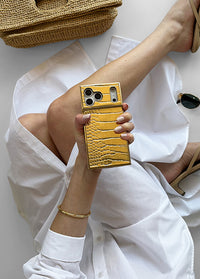 ["Gold", "textured", "iPhone", "case", "with", "crocodile", "pattern", "held", "in", "hand,", "showing", "dual", "camera", "cutout", "and", "square", "shape", "design."]